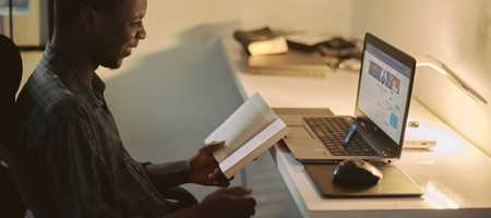 A man at his desk, reading a book whilst smiling in front of his laptops blurred out screen.